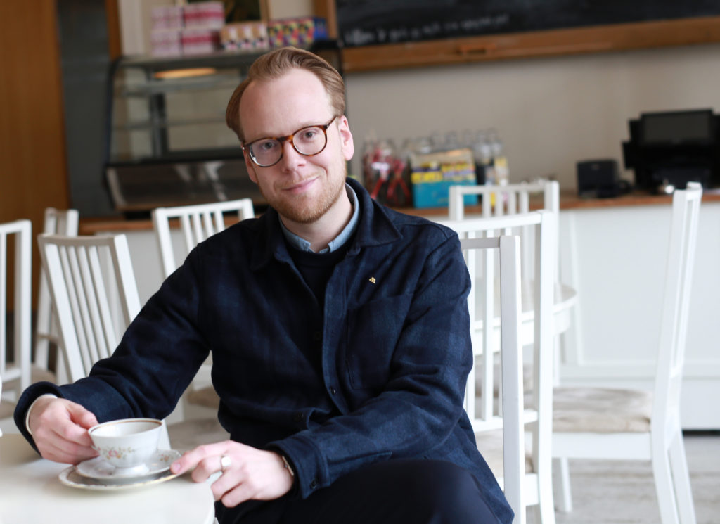 Tobias Pettersson sitter på ett kafe med en kopp kaffe i handen och ser in i kameran.