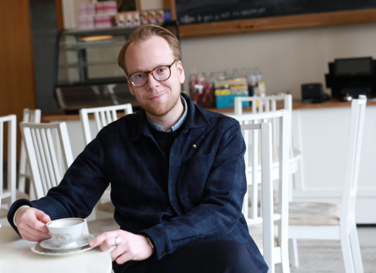 Tobias Pettersson sitter på ett kafe med en kopp kaffe i handen och ser in i kameran.