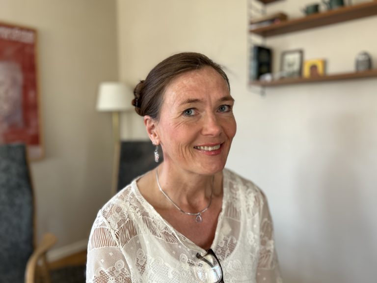 Porträttbild av Jenny Anne sitter hemma i sitt kök och tittar in i kameran och ler.